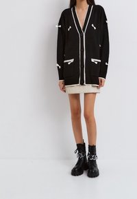 Cardigan en maille noire avec bordure blanche, ornée de nœuds et de poches, fermeture à boutons, et coupe décontractée. Porté sur une jupe claire.