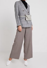 Henkilö, jolla on päällään harmaa ruudullinen tuplaneulostakki, ruudulliset leveälahkeiset housut, valkoiset kärkitossut ja vaaleanvärinen vyölaukku vyötäröllä.