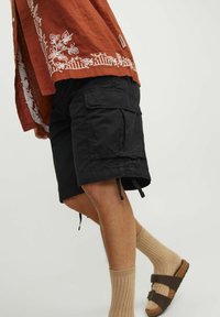 Zwarte cargoshorts met meerdere zakken, vochtige zoom met koord, gedragen met beige geribde sokken en bruine sandalen, gecombineerd met een geborduurd rood shirt.
