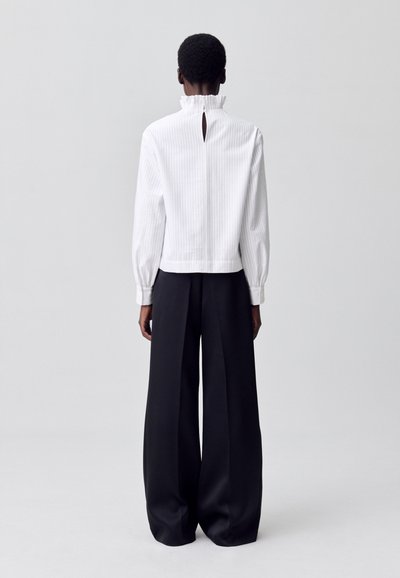Una modelo se encuentra de espaldas al espectador, vistiendo una blusa blanca a rayas con un cuello rizado y pantalones anchos negros, todo ello frente a un fondo claro.