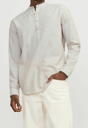 Homme portant une chemise Henley beige clair à manches longues avec des boutons et un pantalon blanc, une main dans la poche, sur un fond uni clair.