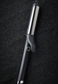 Fer à friser en céramique avec un manche noir et un bouton d'alimentation, reposant sur une surface noire texturée.