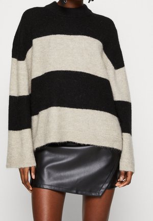 Persona che indossa un maglione a righe larghe nero e beige abbinato a una minigonna in pelle nera su uno sfondo semplice.