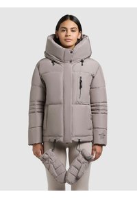Ung kvinde iført en beige polstret jakke med høj krave og aftagelige vanter hængende fra snore, mod en ensfarvet baggrund.