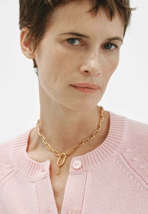 Mujer con cabello corto y castaño, vistiendo un suéter rosa de punto y un collar grueso de cadena dorada con detalle de cierre.