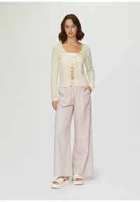 Cremfarbener Strickcardigan mit einem vorderen Bindedetail, kombiniert mit rosa gestreiften weit geschnittenen Hosen. Das Model trägt weiße Plateausandalen.