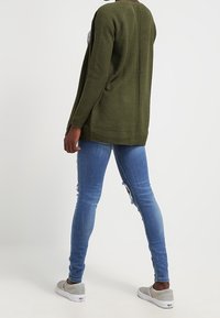 Oliivinvihreä teksturoitu neuletakki rentoa istuvuutta, yhdistettynä vaaleansinisiin kulutettuihin slim fit -farkkuihin ja harmaisiin liukuesteettömiin kenkiin.