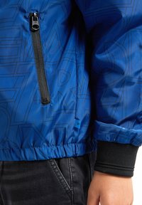 Jachetă bomber albastră cu un finisaj lucios, modele geometrice negre, un buzunar cu fermoar și manșete ondulate. Detaliu de talie înfășurată vizibil.