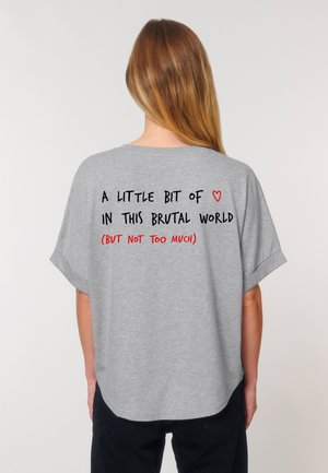 A woman with long hair wears a grey shirt displaying a playful quote about love in a brutal world, emphasizing humor and warmth.