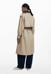 Manteau beige avec un col noir, taille ceinturée et épaules structurées, se caractérisant par des manches longues et un design mi-long.