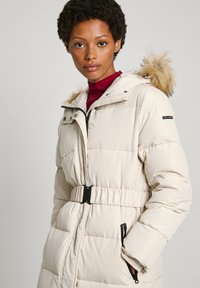 Beige puffjacka med en figursydd design, huva med pälsdetaljer, bälte i midjan och fickor med dragkedja. Slät textur och kviltat mönster.