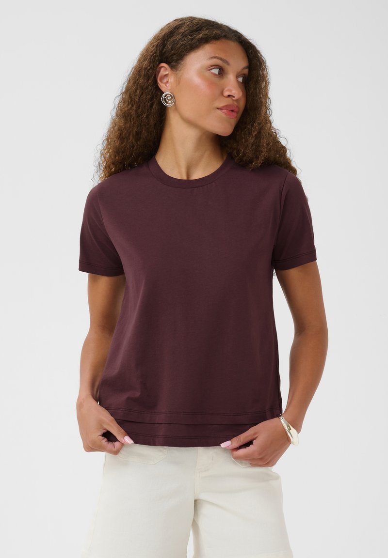 Femme aux cheveux bouclés portant un t-shirt court violet foncé et un pantalon blanc, regardant sur le côté, mains près de l'ourlet du t-shirt.