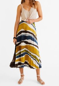 Falda plisada amarilla, azul y marrón con patrones ondulados; combinada con una blusa beige y sandalias de tiras, acompañada de un bolso trenzado.
