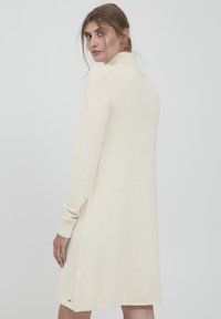 Abito lavorato a maglia di colore crema con colletto alto, maniche lunghe e spacchi laterali. Presenta una texture morbida e una piccola etichetta con il marchio all'orlo.