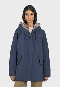 Marineblauwe jas, waterdicht materiaal, grote capuchon met nepbont voering, ritszakken en een relaxte pasvorm voor comfort.