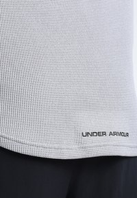 Primer plano de una tela texturizada de color gris claro con el logo negro "Under Armour" cerca del dobladillo, combinado con pantalones oscuros.