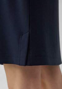 Marineblaue Rockkante mit seitlichem Schlitz, der einen Teil der nackten Beine einer Person vor einem schlichten Hintergrund zeigt.