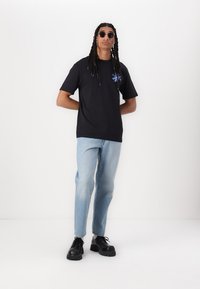 Sort kortærmet T-shirt med et blåt blomsterdesign på venstre bryst, kombineret med lyseblå jeans og sorte platform sko.