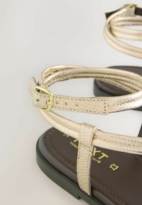 Nærbillede af guldfarvet metallisk ankelremssandal med spænde på mørk sål, der viser tekstureret læder og syningsdetaljer.