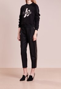 Felpa nera cropped con dettaglio del logo bianco, abbinata a pantaloni neri a sigaretta con una striscia laterale dorata, indossata con scarpe col tacco nere.