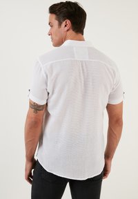 Camisa blanca de manga corta en tejido ligero con un acabado texturizado. Presenta un collar puntiagudo y acentos de botones negros en las mangas.