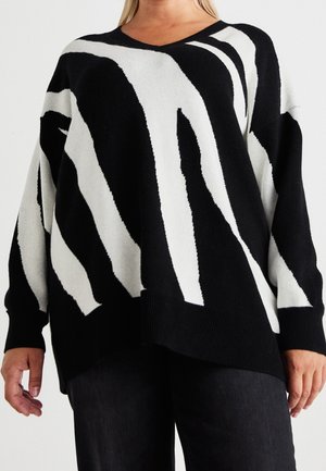 Femme portant un pull oversize noir et blanc avec des rayures abstraites semblables à celles d’un zèbre et un pantalon noir sur un fond clair uni.