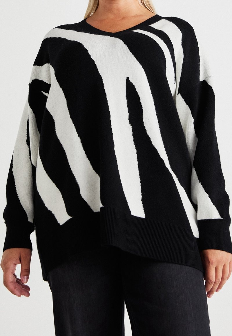 Femme portant un pull oversize noir et blanc avec des rayures abstraites semblables à celles d’un zèbre et un pantalon noir sur un fond clair uni.