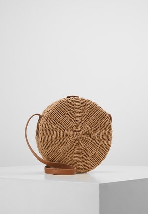 Ronde gevlochten strooien crossbodytas met bruine leren riem, rustend op een wit voetstuk tegen een effen achtergrond.