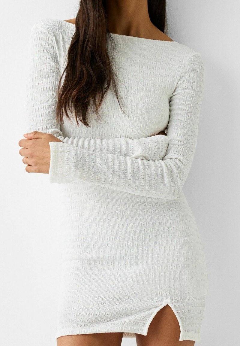 Mini-robe blanche avec un motif côtelé texturé, encolure décolletée épaules nues, manches longues et une fente latérale à l'ourlet.