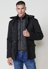 Cappotto nero con chiusura a zip frontale, multiple bottoni e cappuccio. All'interno presenta un rivestimento trapuntato. Indossato sopra jeans blu.