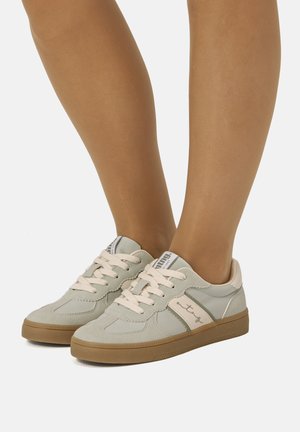 Zapatillas deportivas de cordones en gris claro y beige con suelas de goma, llevadas en piernas bronceadas, mostradas sobre un fondo blanco liso.