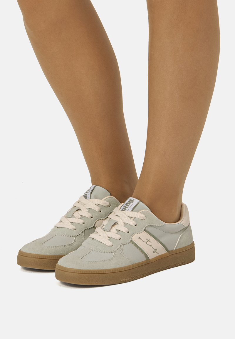 Zapatillas deportivas de cordones en gris claro y beige con suelas de goma, llevadas en piernas bronceadas, mostradas sobre un fondo blanco liso.