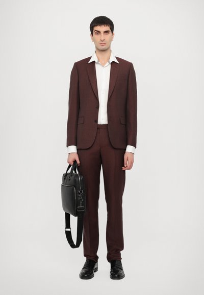 Costume marron avec une veste ajustée et un pantalon, chemise blanche en dessous, chaussures en cuir noir, tenant un sac en cuir noir avec une bandoulière.