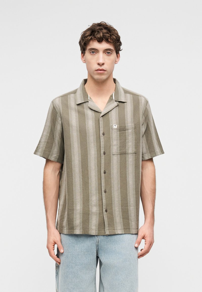 Νεαρός άνδρας που φοράει πουκάμισο με κάθετες ρίγες σε ελιά πράσινο και μπεζ με κοντά μανίκια, κουμπιά και τσέπη στο στήθος, καθώς και ανοιχτόχρωμο μπλε τζιν.