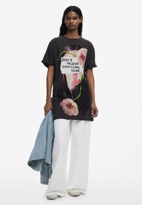 Mustat ylisuuret t-paidat, joissa on kukkakuvio ja teksti "ÄLÄ USKO KAIKKEA, MITÄ AJATTELET", yhdistettynä valkoisiin leveisiin housuihin ja raidalliseen takkiin.