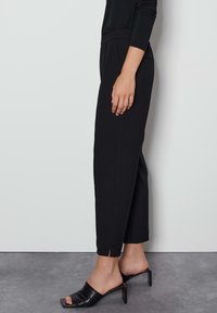 Zwarte op maat gemaakte broek met een gladde textuur, verkorte lengte en zijsplitten aan de zoom, gecombineerd met zwarte hoge hakken.