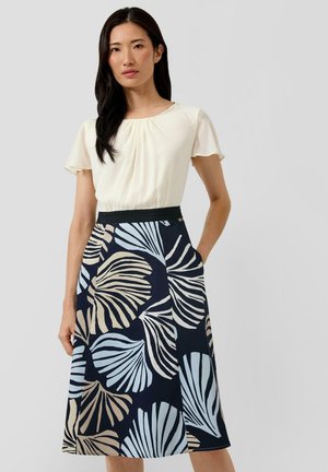 Kobieta ubrana w kremową bluzkę z krótkim rękawem oraz granatową spódnicę z dużymi beżowymi i jasnoniebieskimi wzorami liści, stoi z jedną ręką w kieszeni.