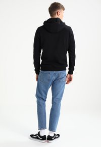 Zwarte hoodie met een kangoeroezak, gedragen met lichtblauwe jeans. De outfit wordt compleet gemaakt met zwarte sneakers en witte sokken.