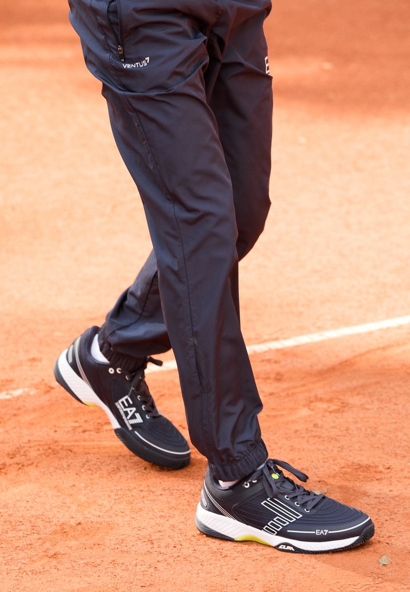 Sapatos de atletismo pretos com detalhes brancos e solas texturizadas, combinados com calças de atletismo azul-marinho, sobre uma superfície de argila avermelhada.