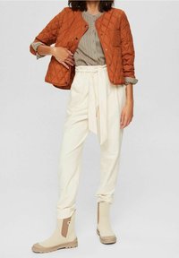 Gesteppten orange Jacke, beiges Hemd, cremefarbene weit geschnittene Hose, die an der Taille gebunden ist, und beige Knöchelstiefel mit braunen Akzenten.