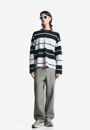 Person wearing white sunglasses, black and white striped long-sleeve shirt, loose gray pants, and gray sneakers, standing against a plain white background.