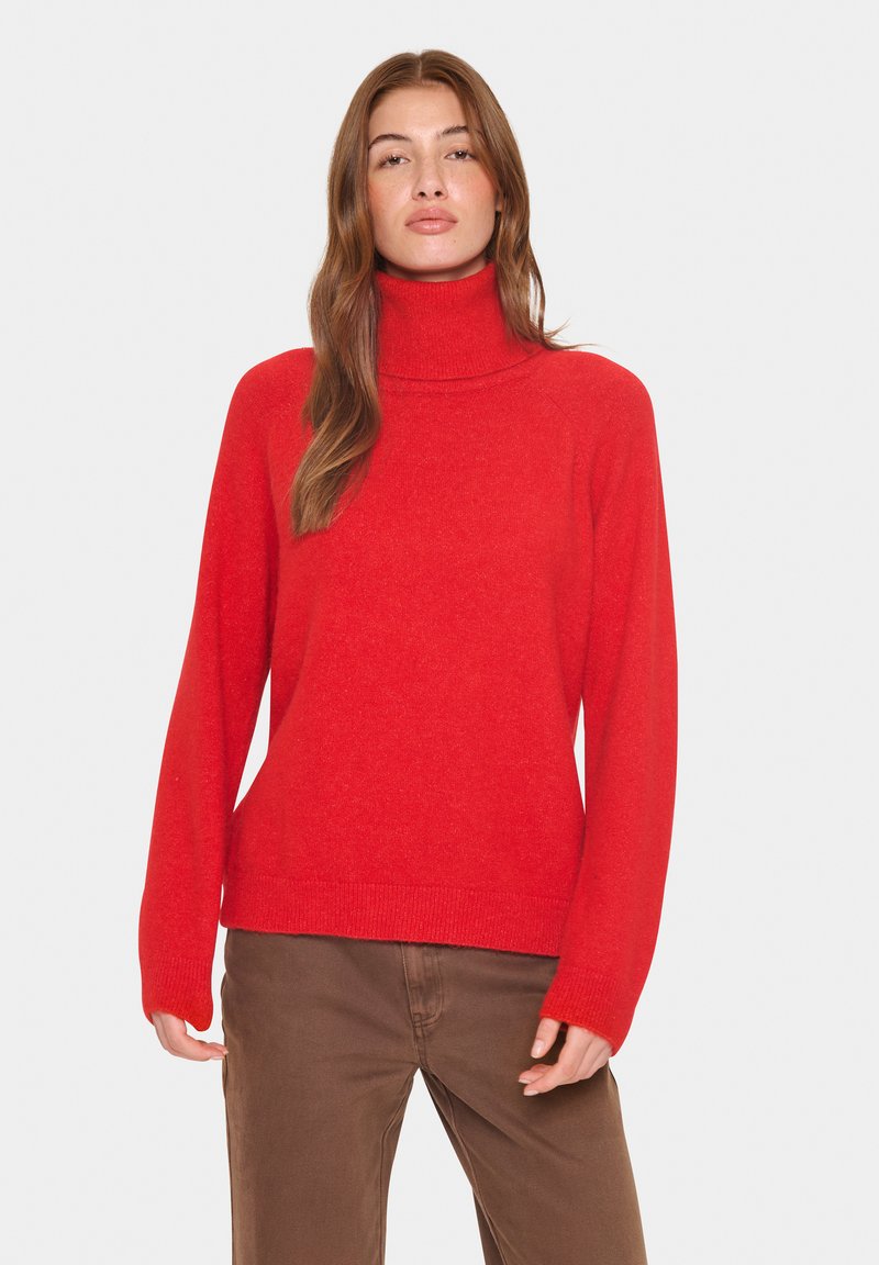 Raudonas nertinis megztinis, pagamintas iš minkštos, tekstūruotos medžiagos, su ilgomis rankovėmis ir laisvu siluetu, derinamas su rudomis kelnėmis.