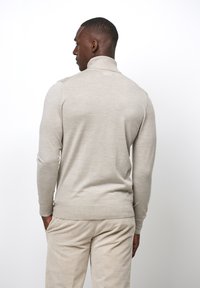 Beige Strickpullover mit hohem Kragen und langen Ärmeln, ausgestattet mit gerippten Bündchen und einem geraden Saum, kombiniert mit strukturierten, hellen Hosen.