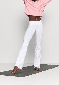 Pantalon de yoga blanc évasé avec une taille haute, associé à un sweat à capuche court rose à cordon. Tissu lisse et extensible.