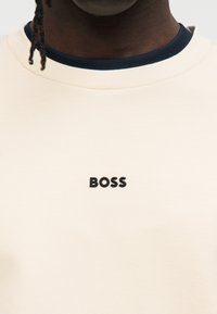 Sweat-shirt beige clair avec un col bleu marine. Présente un logo "BOSS" noir bien visible sur la poitrine. Matière en coton lisse.