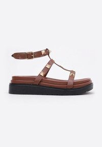Sandales plates marron avec des clous en pyramide dorés, sangle de cheville ajustable et semelle rembourrée à couches. Semelle extérieure en caoutchouc noir pour une bonne adhérence.