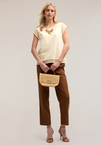 Lichtbeige blouse met korte mouwen, V-hals, gecombineerd met bruine, op maat gemaakte broeken. Houdt een rieten handtas vast met een gouden sluiting en franjes.