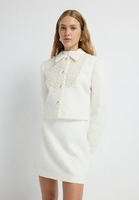 Conjunto de chaqueta y falda blanca con un acabado texturizado. La chaqueta tiene un cuello con cuentas y cierres de botón. Ambas piezas son de longitud a la rodilla.