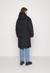 Zwarte pufferjas met een golvende, gequiltte textuur, capuchon en lange mouwen, gecombineerd met lichtblauwe jeans en zwarte chunky laarzen.