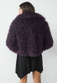 Kręcony, teksturowany fioletowy futerko z luźnym, oversize'owym krojem, z szerokim kołnierzem i opadającymi ramionami, noszone na czarnej sukience.